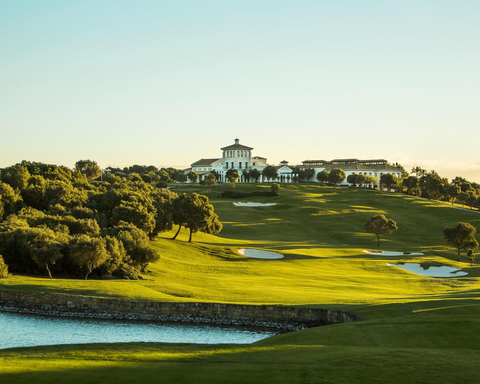 A landscaped golf course with bunkers and a pond at sunset, clubhouse on the hill beyond