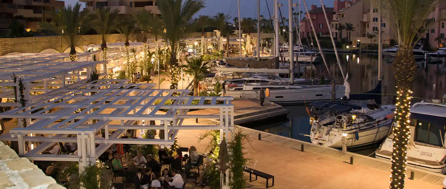 Evening on the marina — a restaurant terrace under a pergola, lit palms and moored sailboats