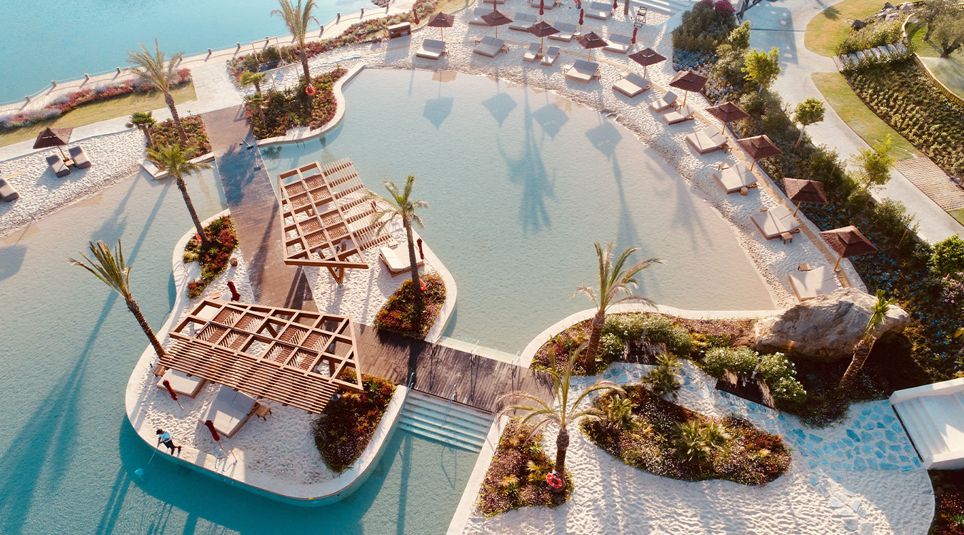 Aerial view of a lagoon-style swimming pool with a sandy beach entry, palm trees and lounge chairs
